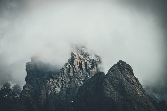Dark atmospheric surreal landscape with dark rocky mountain top in low clouds in gray cloudy sky. Gray low cloud on high pinnacle. High black rock with snow in low clouds. Surrealist gloomy mountains. © Daniil