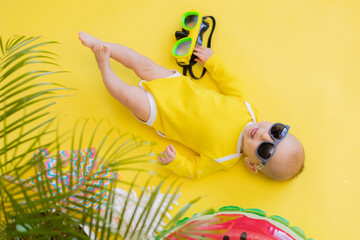 baby in a yellow bodysuit and sunglasses lies under a palm tree next to a bright inflatable lifebuoy and a diving mask. monochrome bright yellow background. summer. space for text. High quality photo