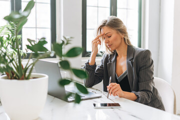 Young tired blonde woman with long hair in stylish grey suit working at laptop in the bright office