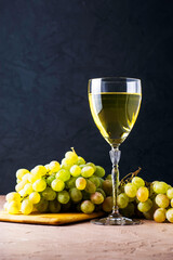 a glass of white wine and a brush of white grapes on a cutting board in the background on a black background