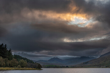 Fototapeta na wymiar Epic stunning Autumn landscape image of Snowdon Massif viewed from shores of Llynnau Mymbyr at sunset with dramatic dark sky and clouds