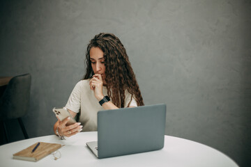 A young brunette girl with long wavy hair in a beige T-shirt works at home with a laptop, holds a...