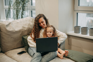A young brunette woman with long wavy hair in jeans and a white sweater sits at home on the sofa in an embrace with her 2-year-old daughter and watches cartoons behind a laptop, next to a smartphone