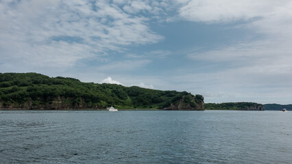 The picturesque coast of Kamchatka. Green hills against a background of blue sky and clouds. A white yacht is sailing across the Pacific Ocean. Avacha Bay.