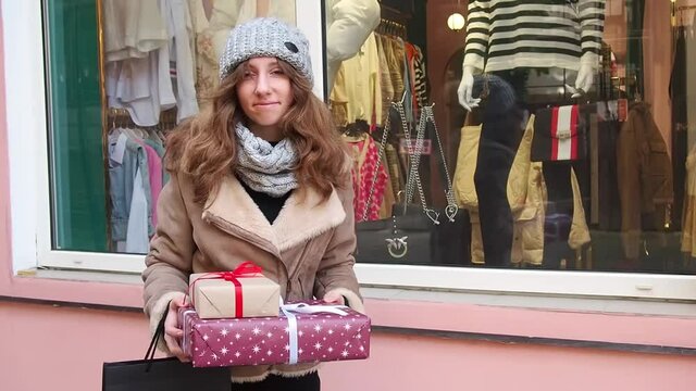 Teengirl in grey hat, scarf and brown jacket holding a set of christmas gifts near shops window. Happy young woman preparing holidays presents. Concept of shopping, giving and receiving gifts.