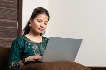 Young indian ethnic girl student freelancer working from home while sitting on the couch, looking at pc screen focused on thinking and solving online problem.