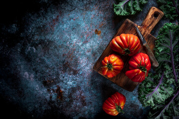 Flat lay of big fresh ripe tomatoes with kale leaves and wooden cutting board on the rustic dark blue background