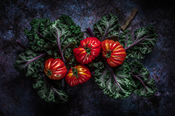 Flat lay of big fresh ripe tomatoes with kale leaves and wooden cutting board on the rustic dark blue background