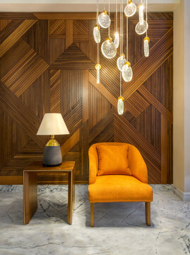 Modern orange armchair, lamp shade on small wooden table, and contemporary tall glass chandelier, in a hall with decorated wood cladding wall, and white marble floor