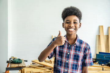 Smiling African-American boy carpenter standing with giving thumbs up as sign of success in a carpentry workshop.