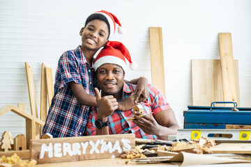 Christmas happy child and father . Cheerful african american son carpenter embracing his father while leaning at the wooden table with diverse working tools laying on it