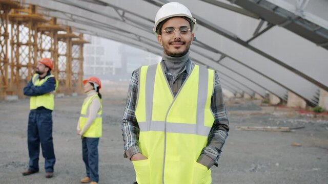 Slow motion portrait of Arab man wearing safety helmet standing in construction site smiling looking at camera while people in uniform working in background