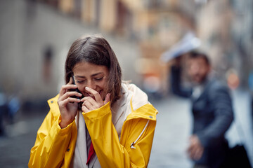 Fototapeta premium A young girl in a yellow raincoat is in a good mood while talking on the smartphone on the street on a rainy day. Walk, rain, city