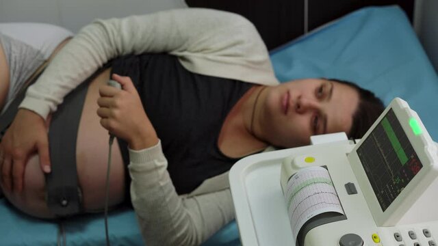 Young Pregnant Woman On Hospital Couch During Medical Control Cardiotocography. Visit Gynecologist Doctor At Clinic For Pregnancy Consultant. Examine Expectant Belly Baby Mother Healthcare Check Up