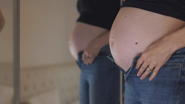 4K footage of a pregnant girl trying to wear a pair of jeans in front of a mirror, but her belly is too big
