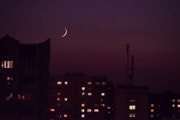 Young crescent moon on the sky over city. Silhouette of towers and houses with chimneys on roofs....