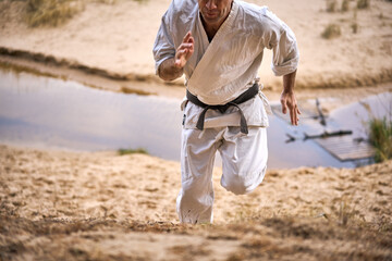 Karate man with black belt in an old kimono runs up a hill of sand. A small river flows below. Concept.