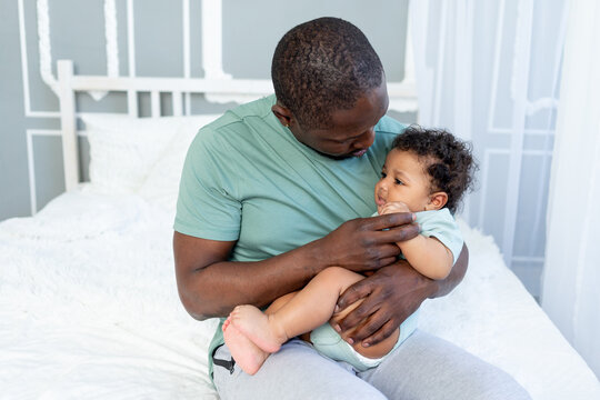 an African-American dad calms or puts his son to sleep by holding him in his arms on the bed at home, a caring father