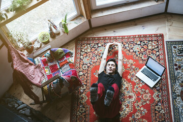 Obraz na płótnie Canvas Young man doing warm-up during a break while working at home. Lying on vintage carpet near laptop and rocking chair