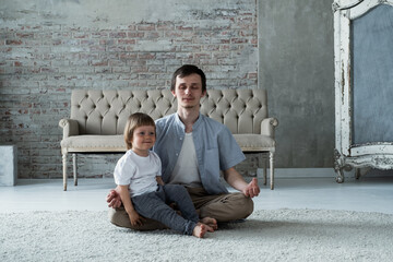 Fototapeta na wymiar Young father trying to meditate sitting in lotus yoga pose with little son kid