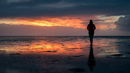 Fototapeta na wymiar Woman silhouette walking along the Wadden Sea in Buesum during sunset, reflecting in the wet sand, Germany.