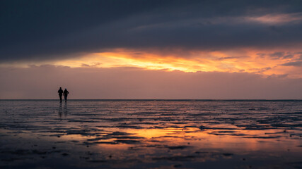 Couple silhouette walking along the Wadden Sea in Buesum during sunset, reflecting in the wet sand,...