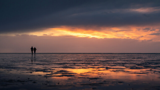 Couple silhouette walking along the Wadden Sea in Buesum during sunset, reflecting in the wet sand, Germany.