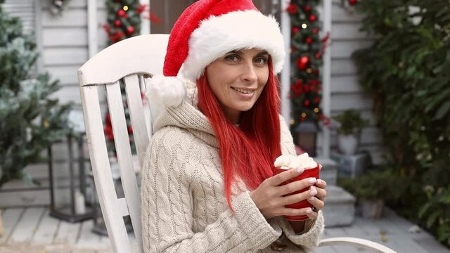 Beautiful hipster girl with red hair in a santa claus hat sits in a rocking chair on the veranda of the house, enjoys the winter holidays and christmas mood and drinks cocoa with marshmallows 