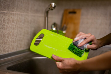 Woman's hands with sponge washing green cutting board in the sink. Housework in the kitchen concept. Dishwasher