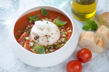 Tomato gazpacho soup served with burrata cheese in a white bowl, studio shot on a light-grey marble background