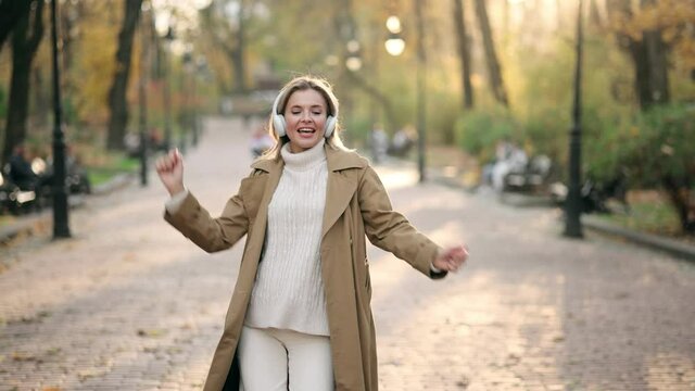 Gorgeous lady in autumn outdoor. Portrait of an attractive adult blonde woman in wireless headphones jumping, dancing and having fun outdoors in the park at fall on a sunny day and smiling