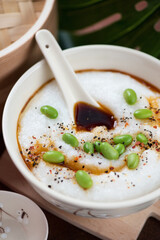 Closeup of congee or rice porridge served with edamame beans and soy sauce in a beige bowl, vertical shot