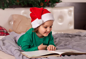 Naklejka premium Little cute girl in Santa cap and Christmas pajamas reading a book.