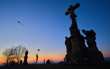 Silhouettes of kids flying kites with stone christianity monument in the foreground at sunset.