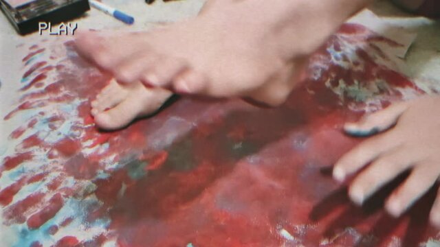 The feet of a kid playing with color paint (drawing on paper with it), recorded on camera with a fake VHS look.
