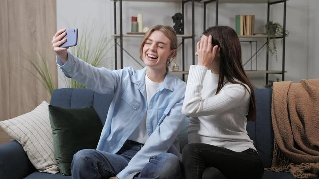 Happy girlfriends talk laugh looking at smartphone using funny apps sit on couch, smiling girls relaxing at home doing selfie having fun in social media on cellphone online watching video on phone.