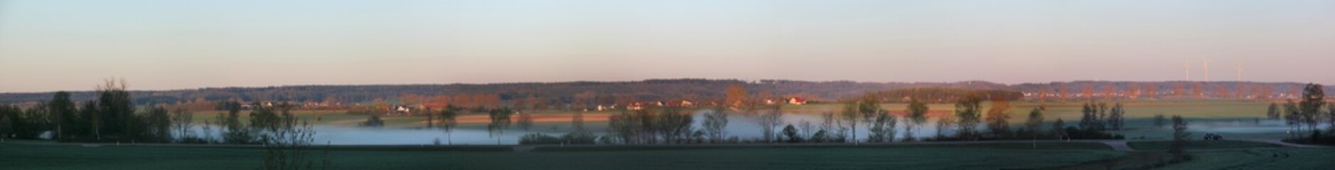 Fototapeta na wymiar Panoramic view of the lowlands of Altmühl river valley on a foggy spring morning with the villages of Stadel, Hilsberg and Aurach in the background, Franken region in Germany