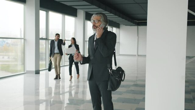Businessman in suit is speaking on mobile phone then talking to builder wearing uniform indoors in modern building. People and communication concept.