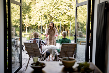 Woman giving cocktails to a friends sitting on wooden chairs at home terrace. European girls and latin man spending time together. Concept of leisure. Modern domestic lifestyle