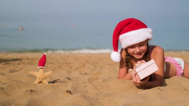 A child lies on the beach wearing a Santa hat. Little girl smiles and looks at a box with a gift. Merry Christmas. Winter vacation. Sea in the background.Copy space.