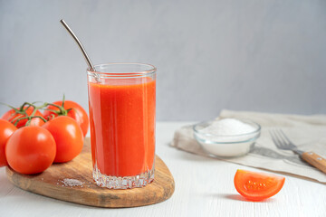 Refreshing homemade tomato juice made from ripe red tomatoes, usually used as a beverage, served in tall drinking glass with reusable metal straw on white wooden table with salt, fork and textile