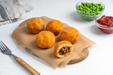 Arancini Italian or Sicilian cuisine rice balls stuffed with minced meat and green peas, coated with bread crumbs and deep fried served on board with fork and ketchup sauce on white wooden table