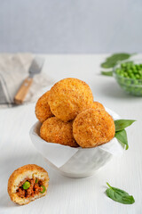 Homemade tasty Arancini Italian or Sicilian rice balls stuffed with minced meat and green peas, coated with bread crumbs and deep fried served in bowl with spinach leaves on white wooden table