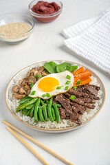 Bibimbap asian or korean rice dish topped with vegetables, sliced beef meat, carrot, mushroom and fried egg with yolk served in plate seasoned with scallions on white wooden table with chopsticks
