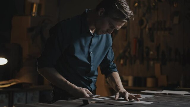Handmade leather production. Professional man tailor cutting fabric on workplace table at workshop in evening, side view