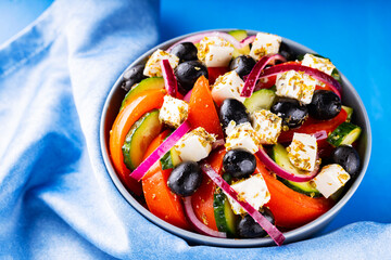 Greek salad and blue napkin on a blue background. Fresh vegetables, feta cheese and black olives. Greek cuisine. Top view. Copy space