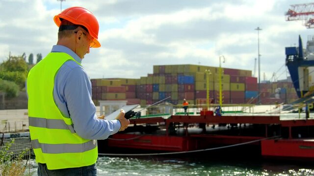 Ship cargo loading process. Safety inspector uniform and helmet marks notes on tablet while worker walk along ferry ramp in dockyard on windy day