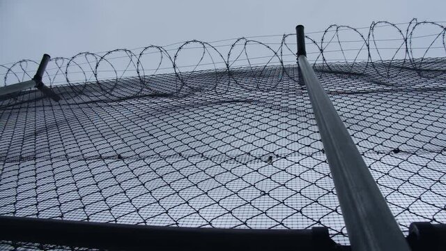 A chain-link fence with barbed wire of the prison or concentration camp with a grey cloudy sky backdrop
