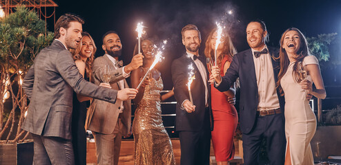 Group of beautiful people in formalwear holding sparklers and smiling while spending time on outdoor party
