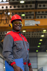 Smiling African American Worker In Protective Workwear Holding Clipboard And Looking At Camera - Portrait Of Black Industrial Worker In Red Helmet, Safety Goggles And Noise Reduction Earmuff